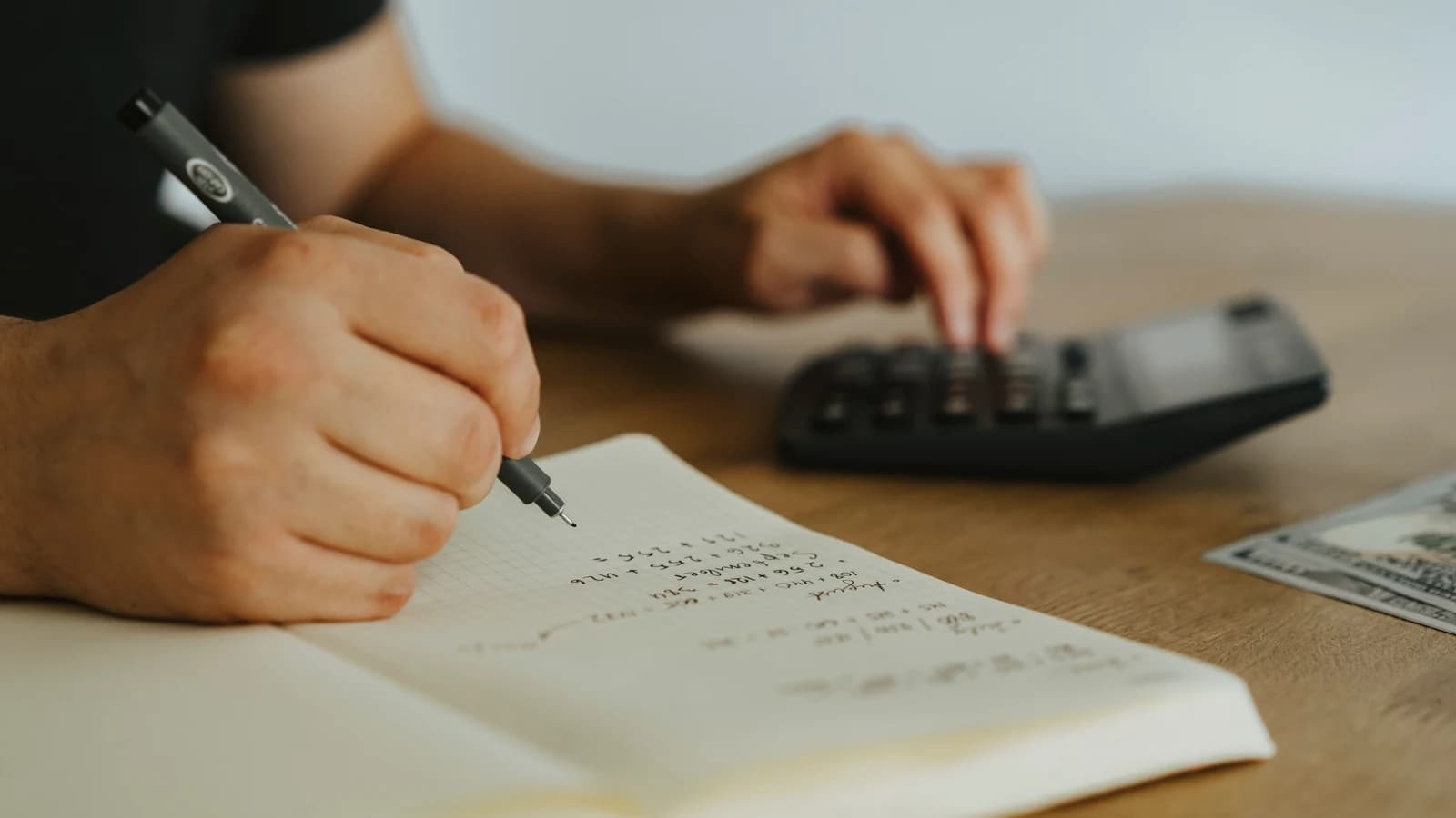Hands writing calculations in a notebook beside a calculator