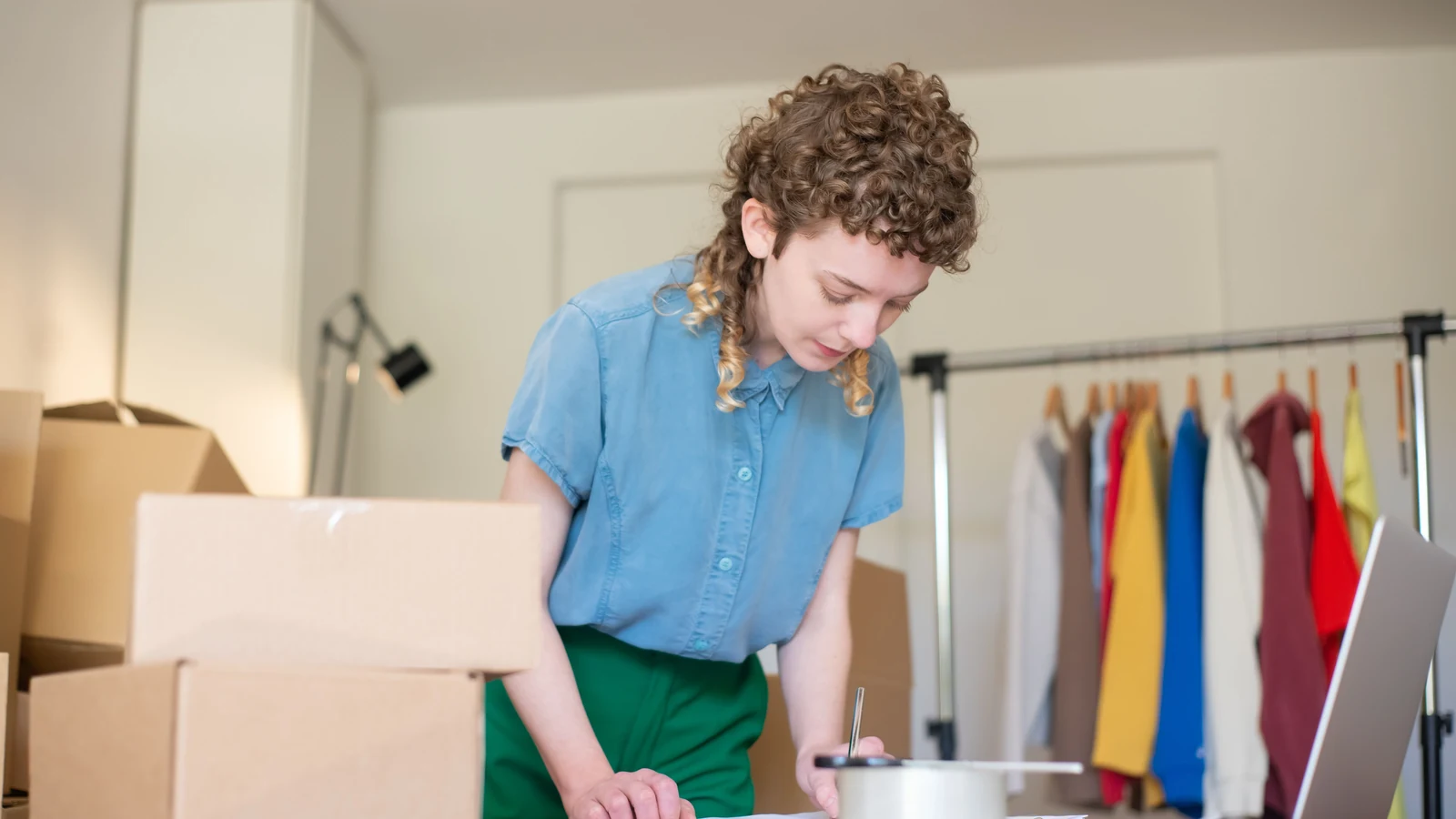 Woman writing beside a laptop and boxes in a home business workspace